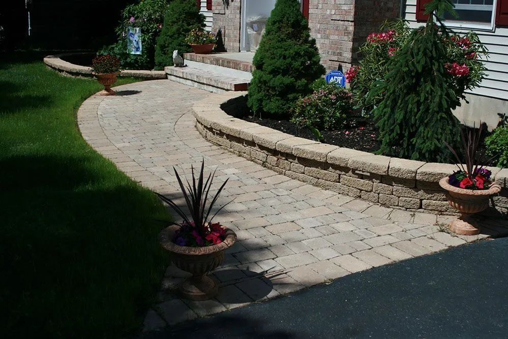 Curved stone garden walkway beside a brick house with potted plants and shrubs in sunlight