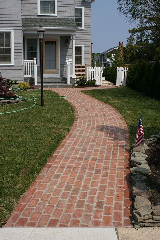 Curved brick walkway leading to a house, bordered by grass, rocks, and a small American flag.