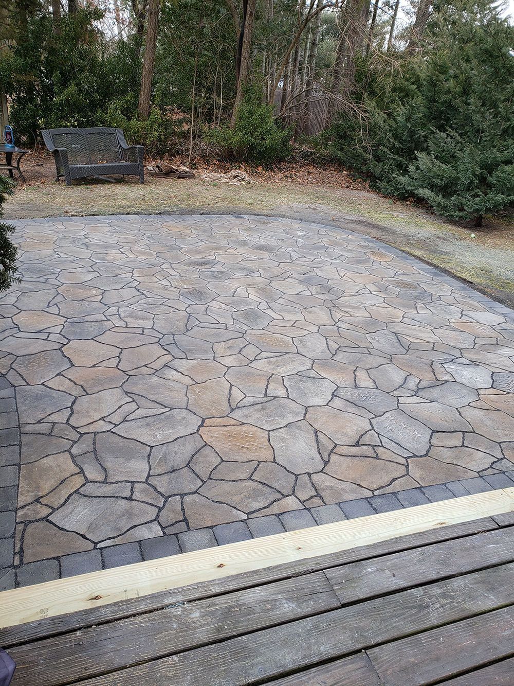 Stone patio with a circular fire pit area, bordered by trees and a wooden deck in the foreground