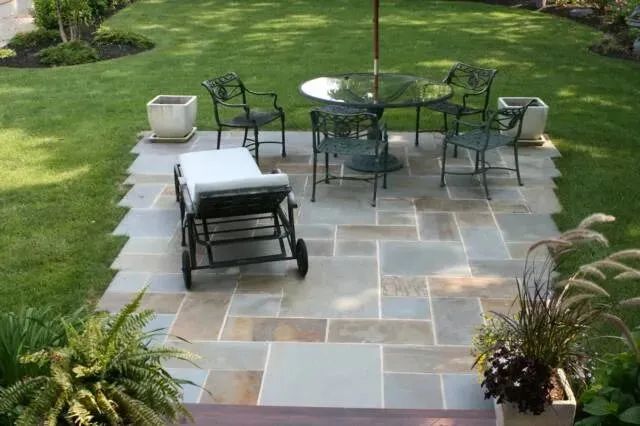 Patio with stone pavers, black metal table and chairs, lounge chair, and potted plants in a grassy yard