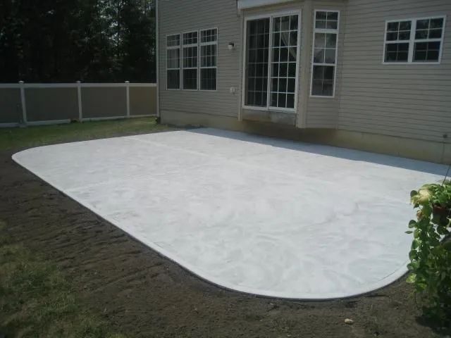White concrete patio beside a beige house, bordered by grass and a fence.