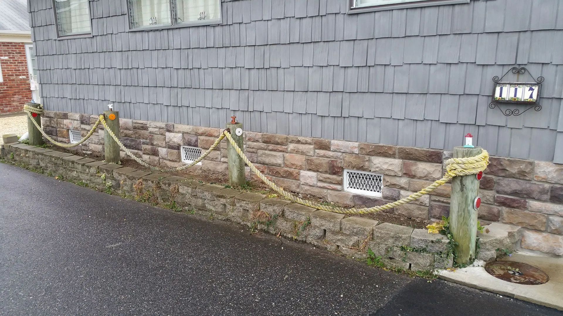 Rainy residential side yard with a low rope fence, stone wall, and gray shingle siding beside a wet path