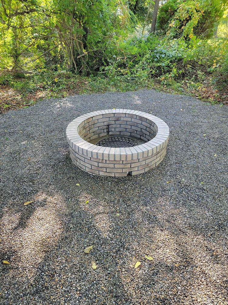 Stone-lined fire pit centered on a gravel clearing with trees and bushes in the background