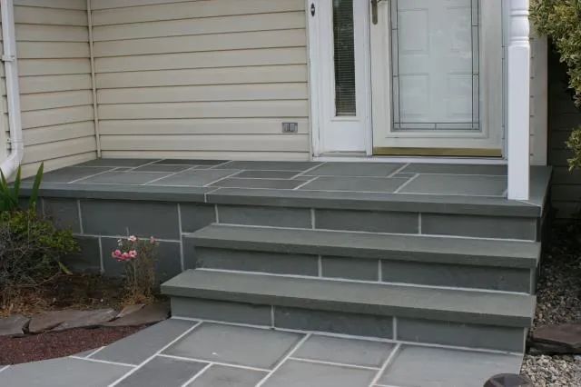 Front porch steps with gray tile pavers and white railing beside a house entrance