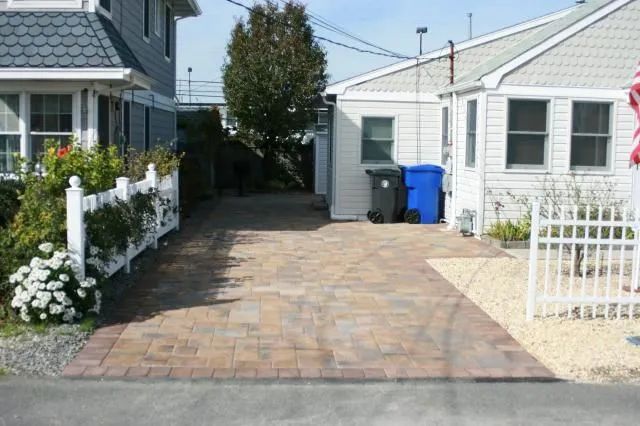 Brick driveway between two houses, with white fences, potted flowers, and blue recycling bins.