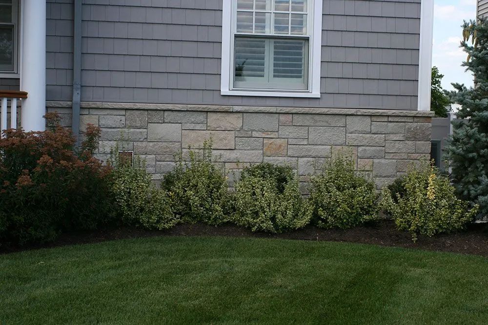 House exterior with gray siding, stone foundation, white window, shrubs, and green lawn