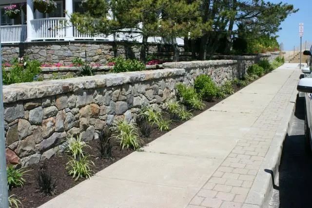 Stone retaining wall beside a paved seaside walkway with flowers and trees on a sunny day