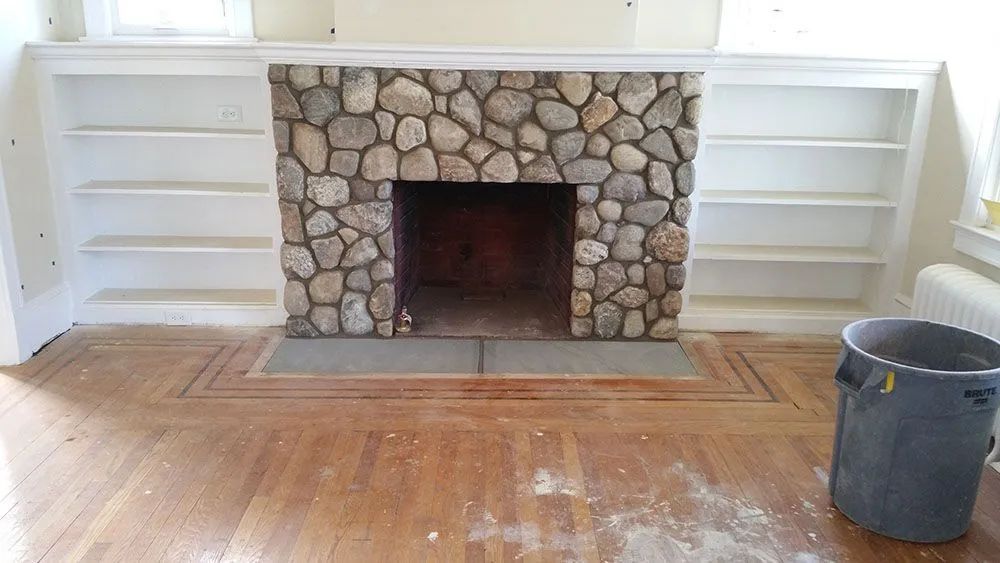 Stone fireplace with white built-in shelves on both sides in an empty room, bucket on wood floor