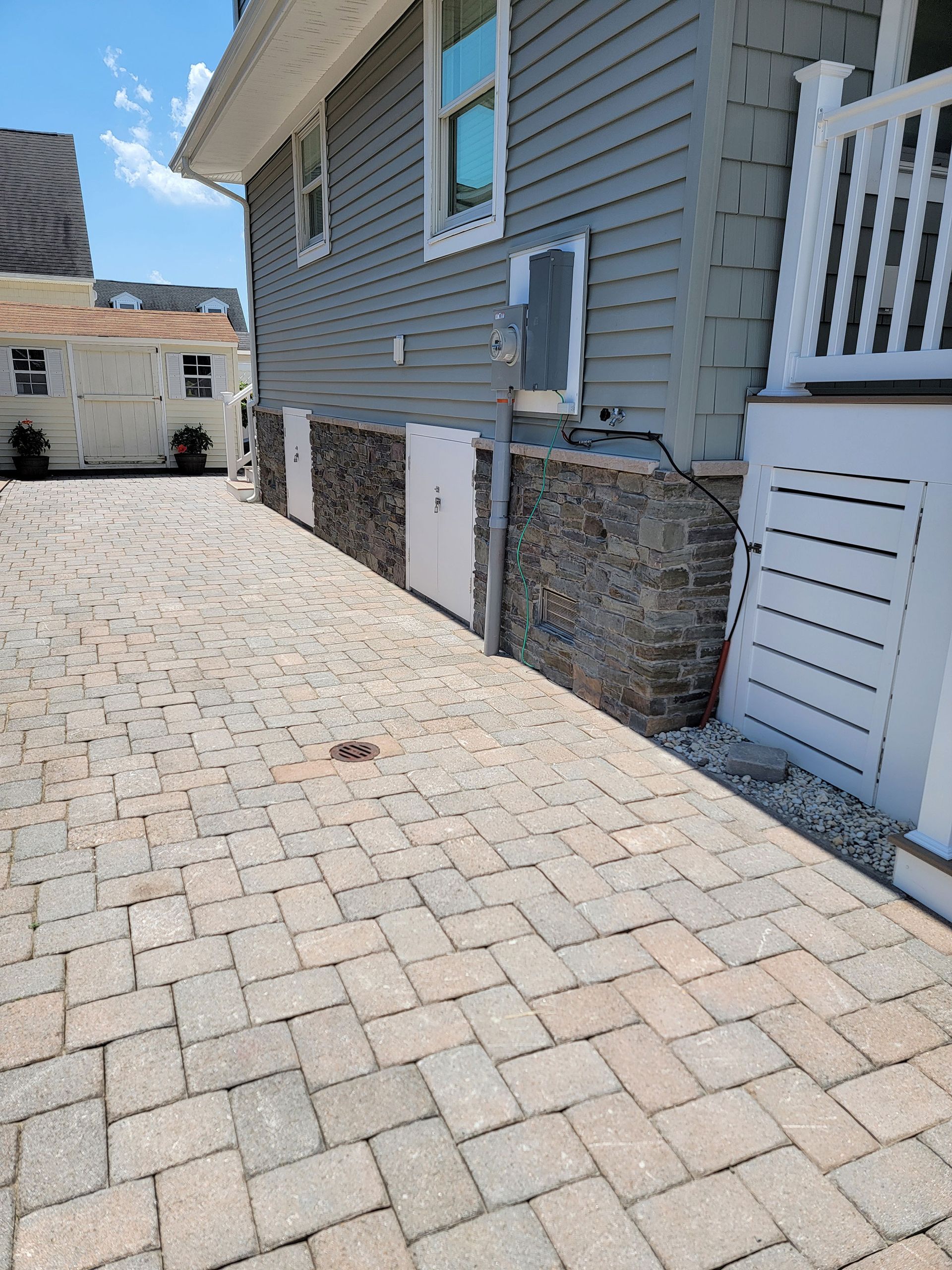 Side yard with cobblestone paving beside a gray house and white porch railing under a blue sky