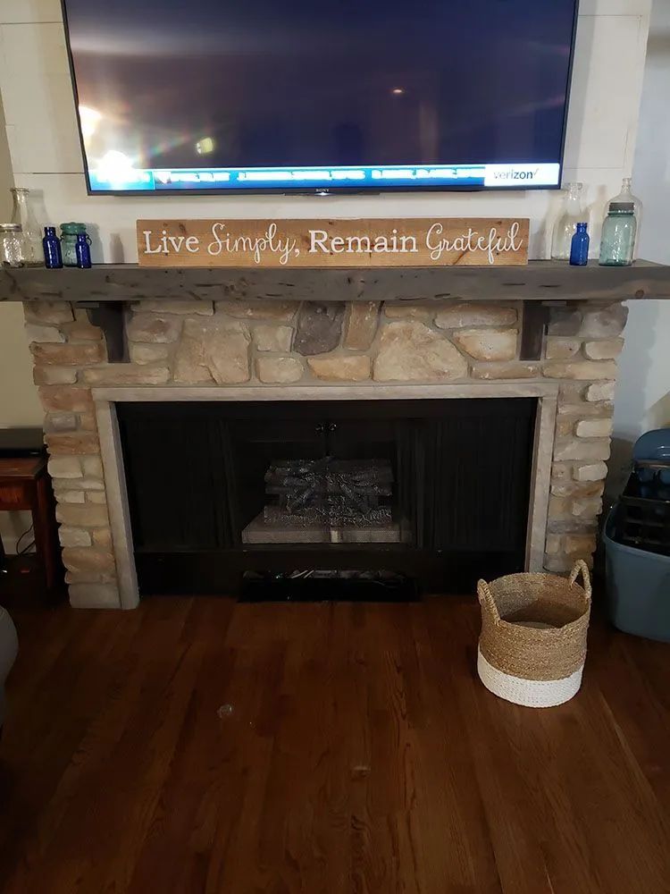 Living room fireplace with TV above, stone mantel, and a “Live Simply, Remain Grateful” sign.