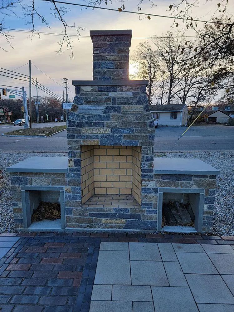 Outdoor stone fireplace with a chimney and built-in benches on a paved patio at dusk