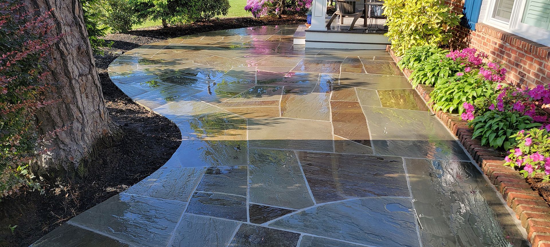 Wet stone walkway through a landscaped garden with pink flowers and trees reflecting in puddles