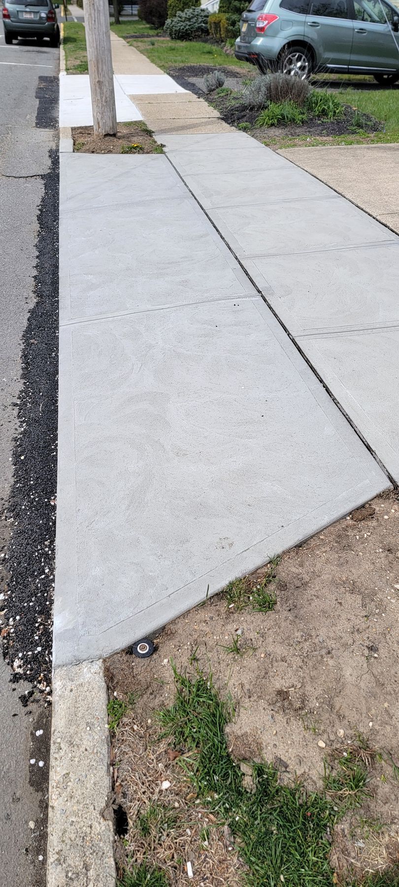 Cracked sidewalk slab with a raised section beside a curb and parked cars.
