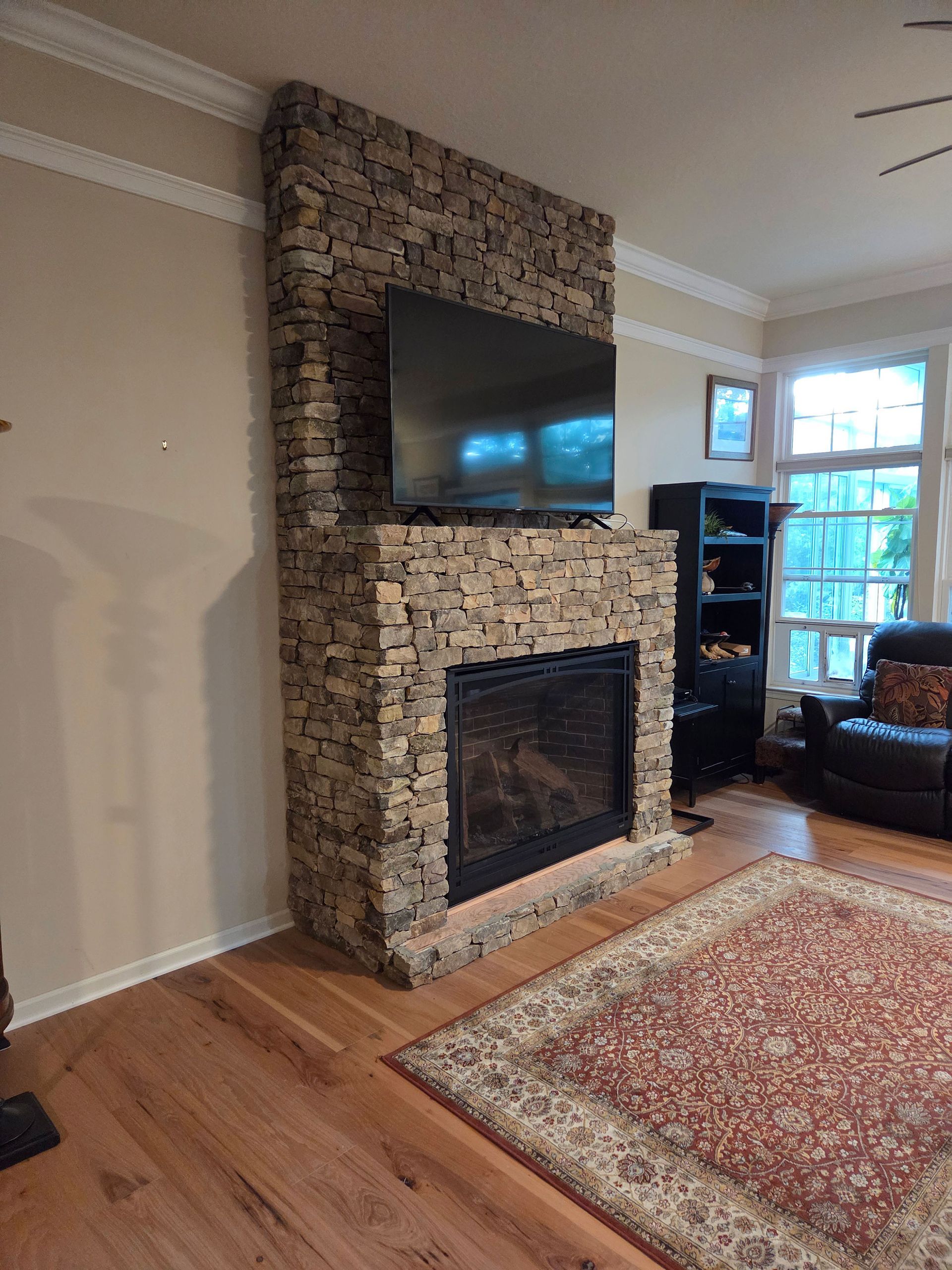 Living room with stone fireplace, wall-mounted TV, rug, hardwood floors, and a large window.