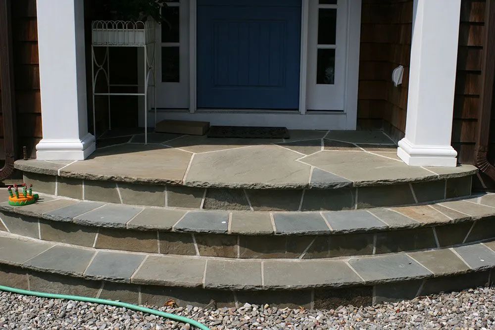 Front porch with blue door, white columns, and curved stone steps leading up from a gravel path.