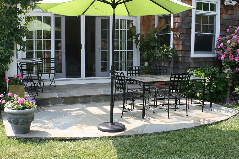Patio with green umbrella, metal table and chairs, and potted flowers outside a house