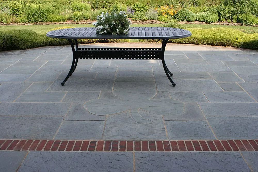 Black metal patio table centered on a gray stone terrace with garden greenery in the background