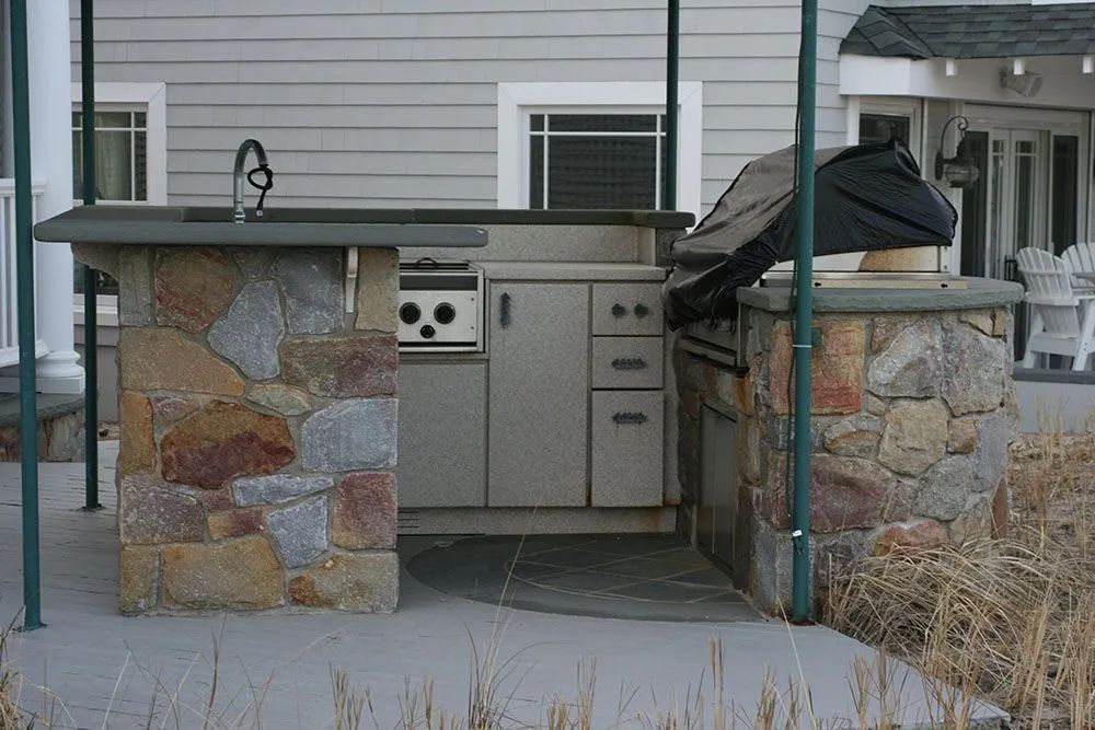 Stone outdoor kitchen with grill, cabinets, and black cover between two stone pillars on a patio.