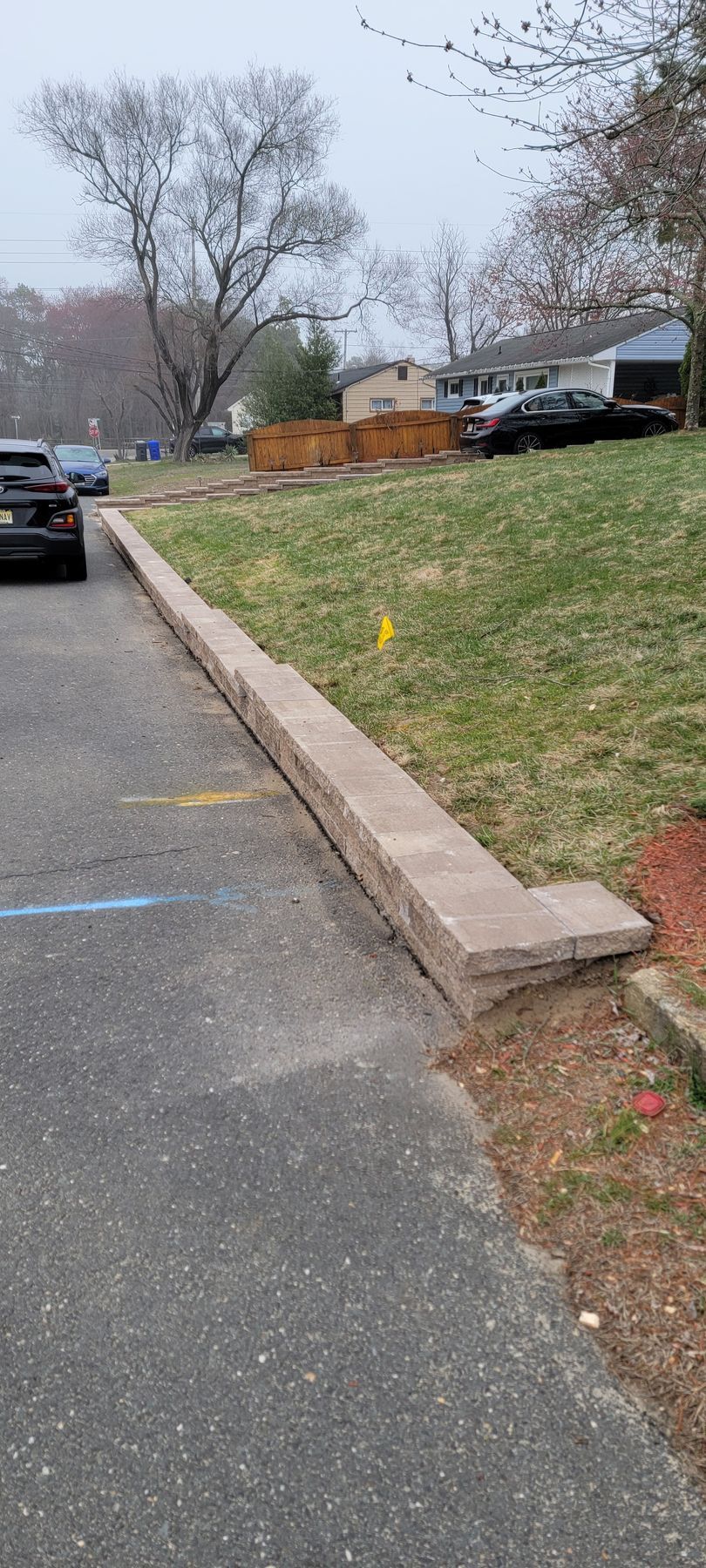 Parking lot with a low stone retaining wall and grassy slope beside trees and parked cars