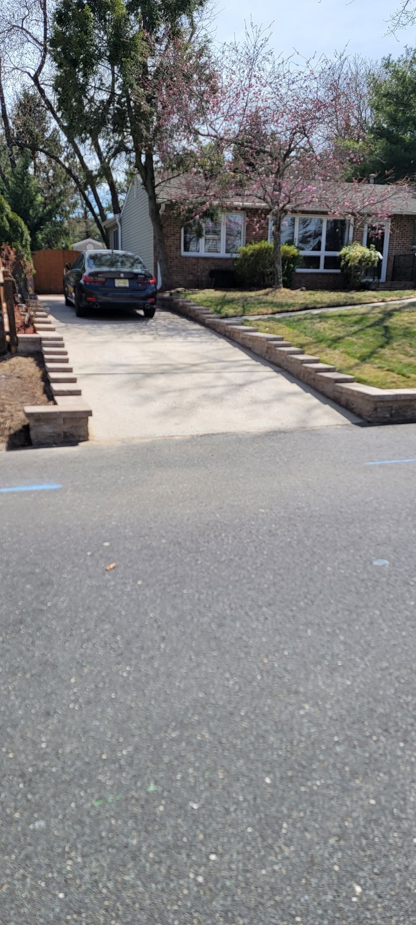 Driveway leading to a house with a parked black car, brick retaining walls, and flowering trees.