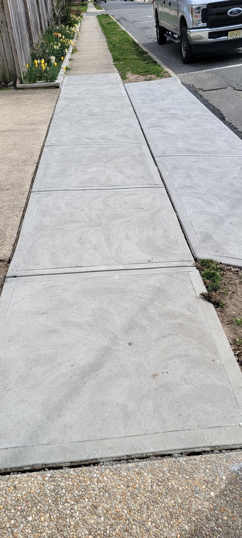Sidewalk with cracked concrete slabs beside a parked car and a narrow strip of grass.
