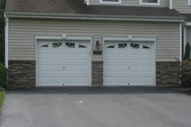 Two white garage doors on a beige house with stone accents and a paved driveway