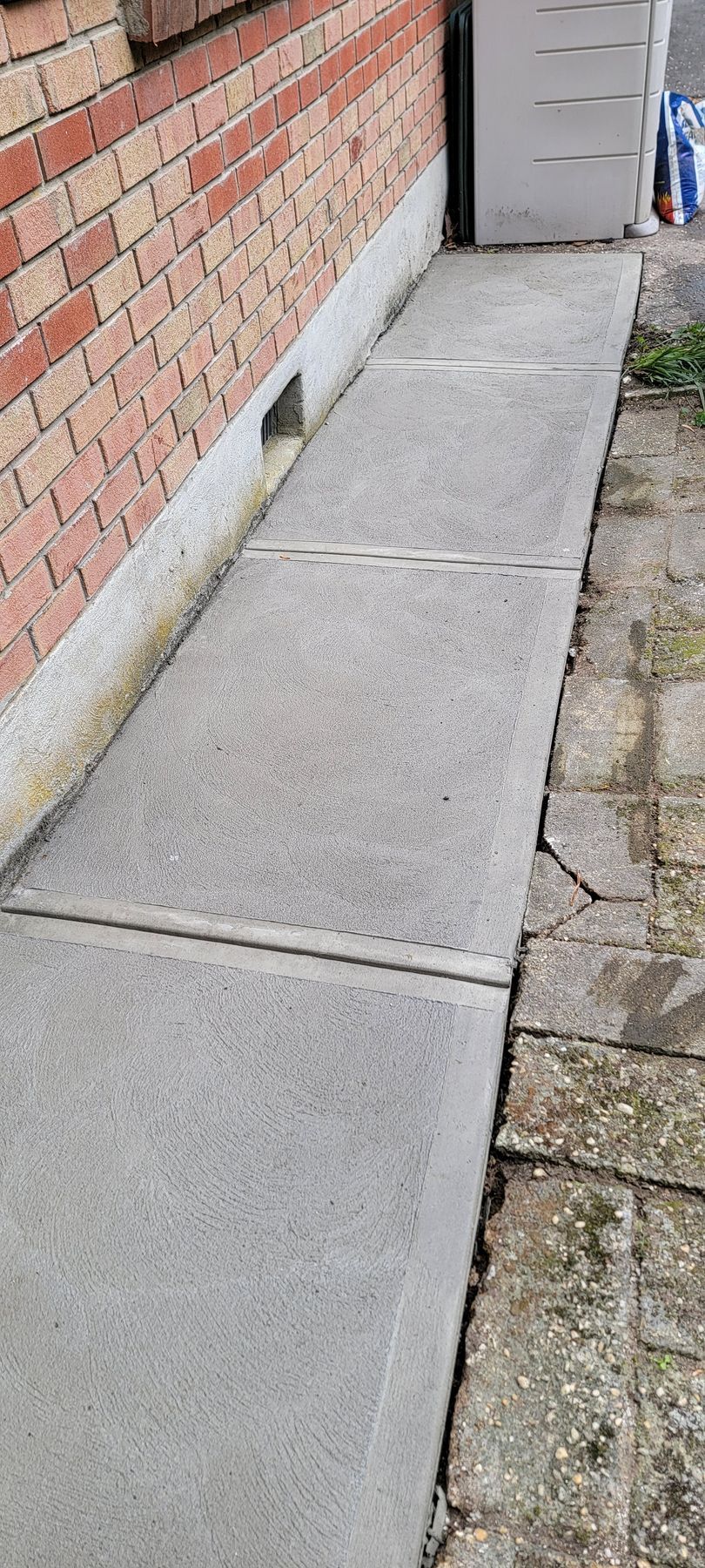Concrete sidewalk beside a red brick wall, with a utility box and cobblestone edge along the path