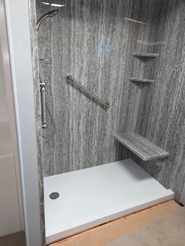 A modern walk-in shower with gray marbled walls, a white floor, a grab bar, a built-in seat, and corner shelves.