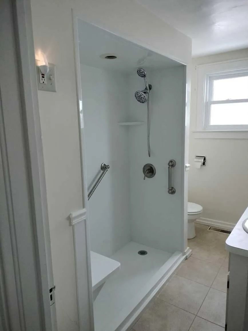 A white walk-in shower with a built-in seat, grab bars, and adjustable handheld showerhead in a modern bathroom.