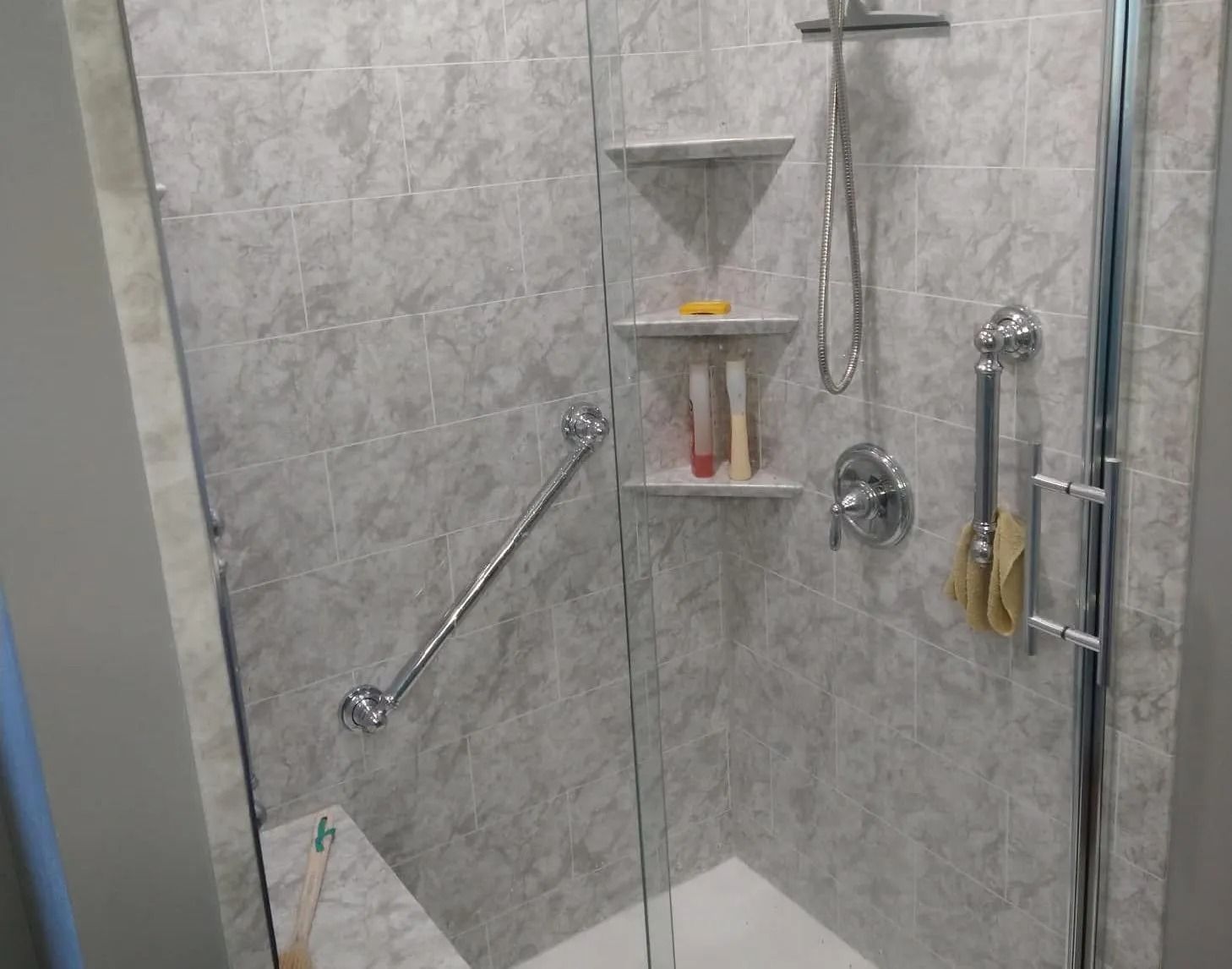 A walk-in shower with gray tiled walls, a glass door, a built-in bench, corner shelves, a handheld sprayer, and grab bars.