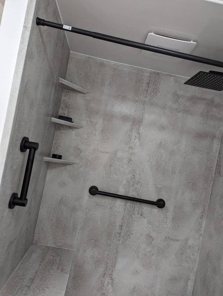 A grey, tiled walk-in shower featuring matte black grab bars, a shower rod, and built-in corner shelves.