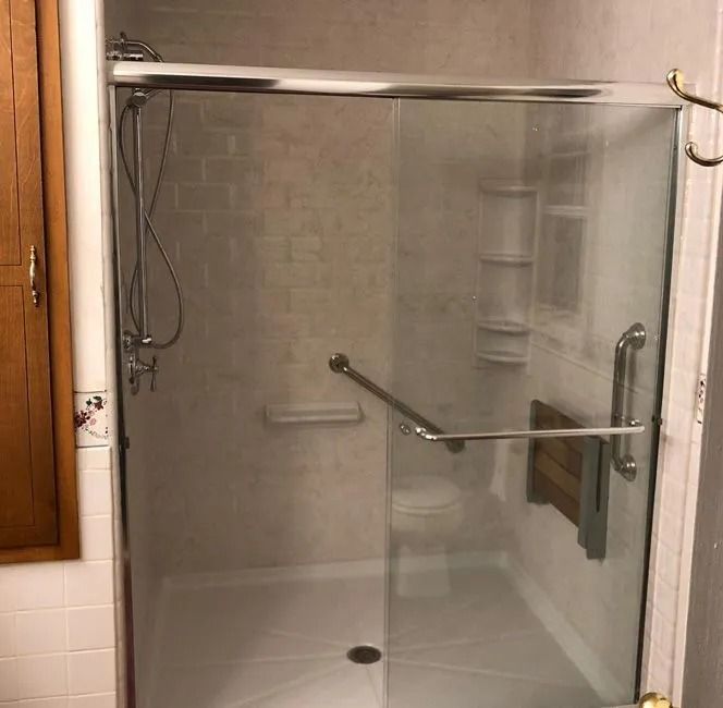 A walk-in shower with a glass sliding door, metal grab bars, a folding wooden seat, and white tiled walls.