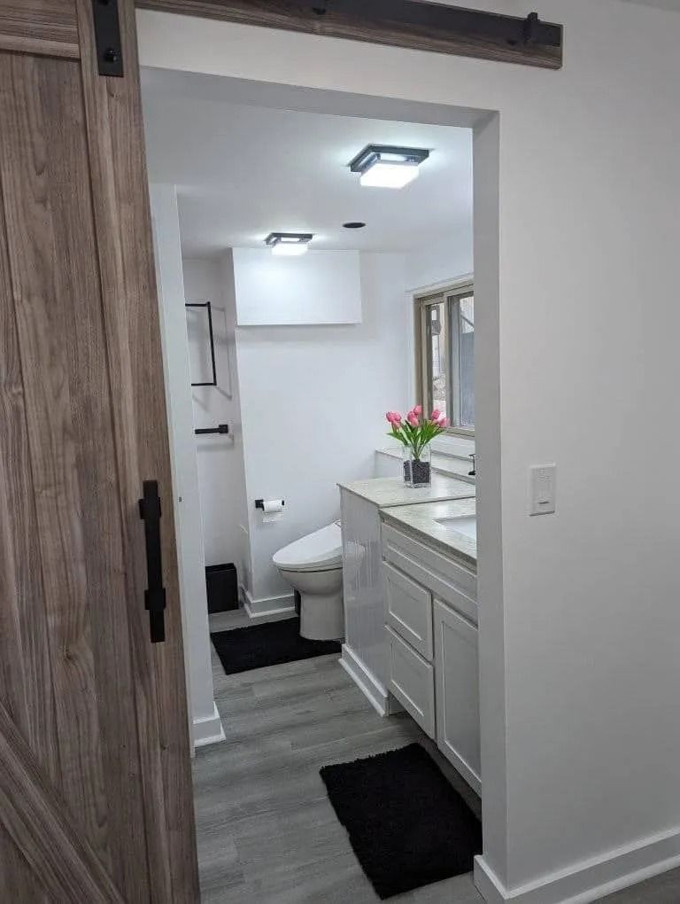 A bright, modern bathroom seen through a rustic sliding barn door, featuring white cabinets, a toilet, and gray flooring.