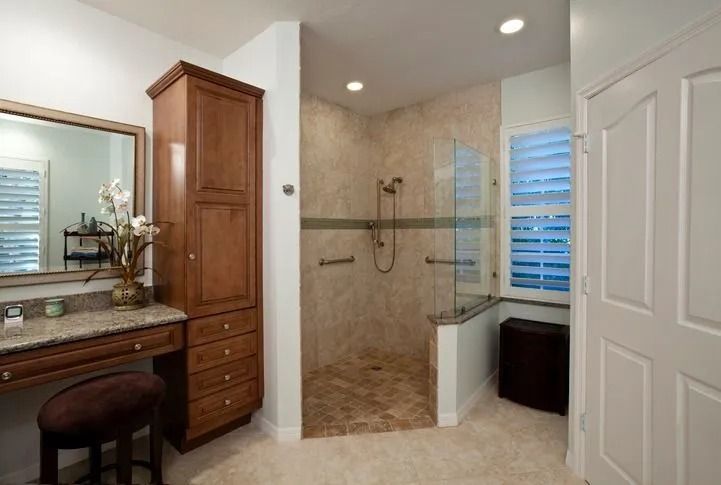 A bathroom vanity with a wooden cabinet and stool next to a walk-in tiled shower with a glass partition and shutters.