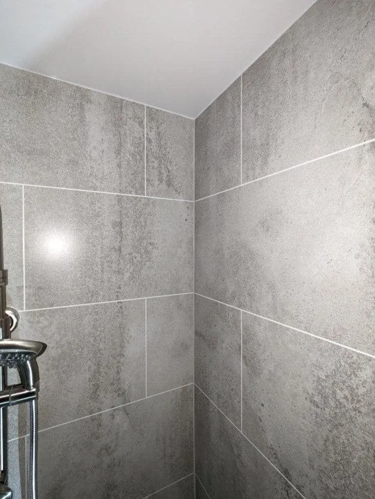 A shower corner with gray, stone-textured rectangular wall tiles and a chrome shower head fixture on the left.
