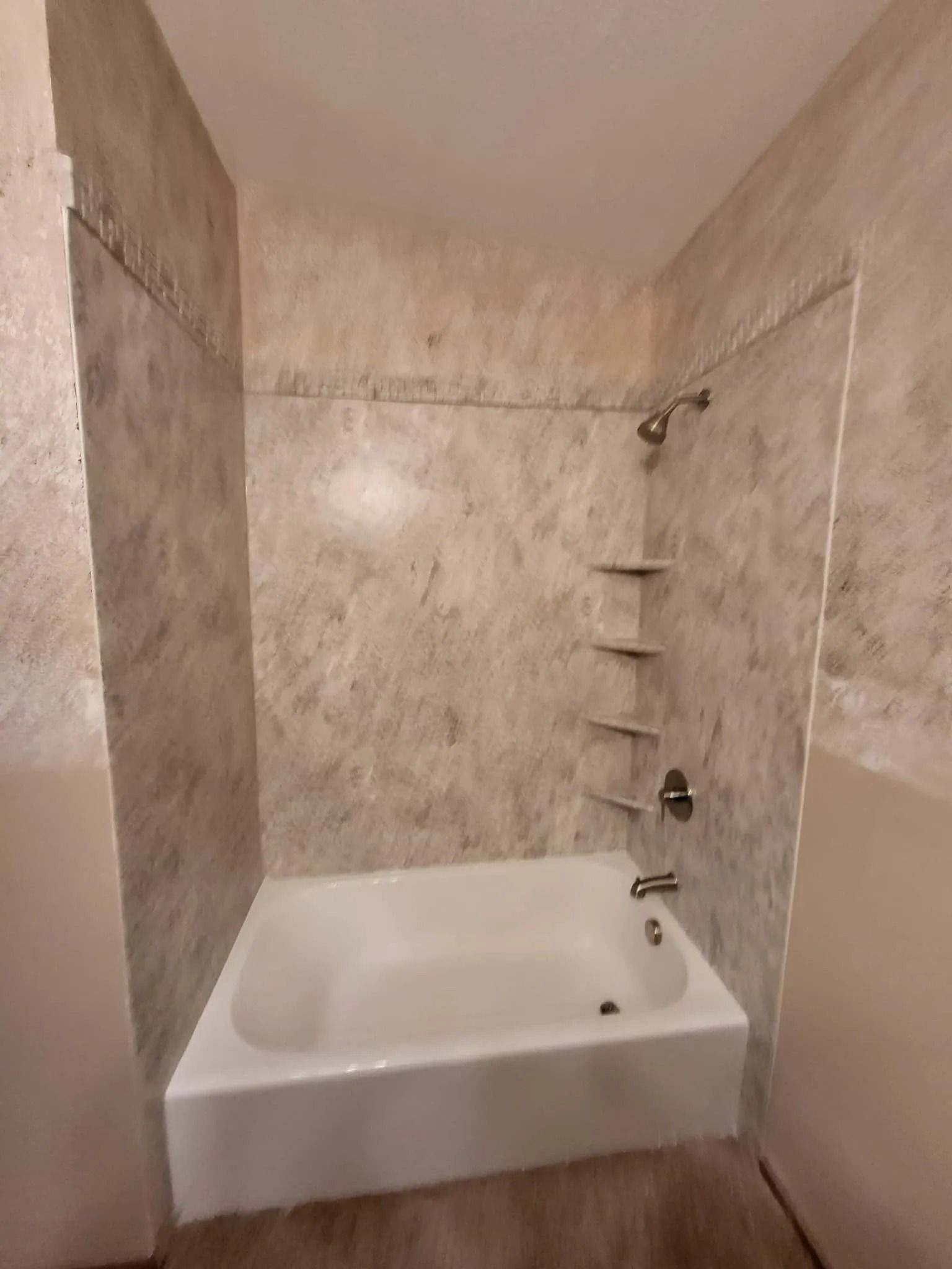 A white bathtub with a shower fixture and built-in corner shelves against marbled tile walls in a bathroom.