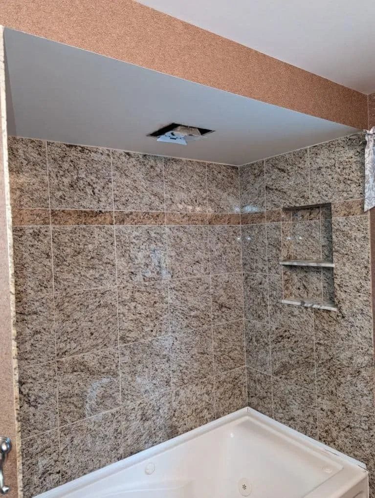A bathtub shower stall with tan granite-patterned tile walls and a built-in shelf, featuring a light fixture in the ceiling.