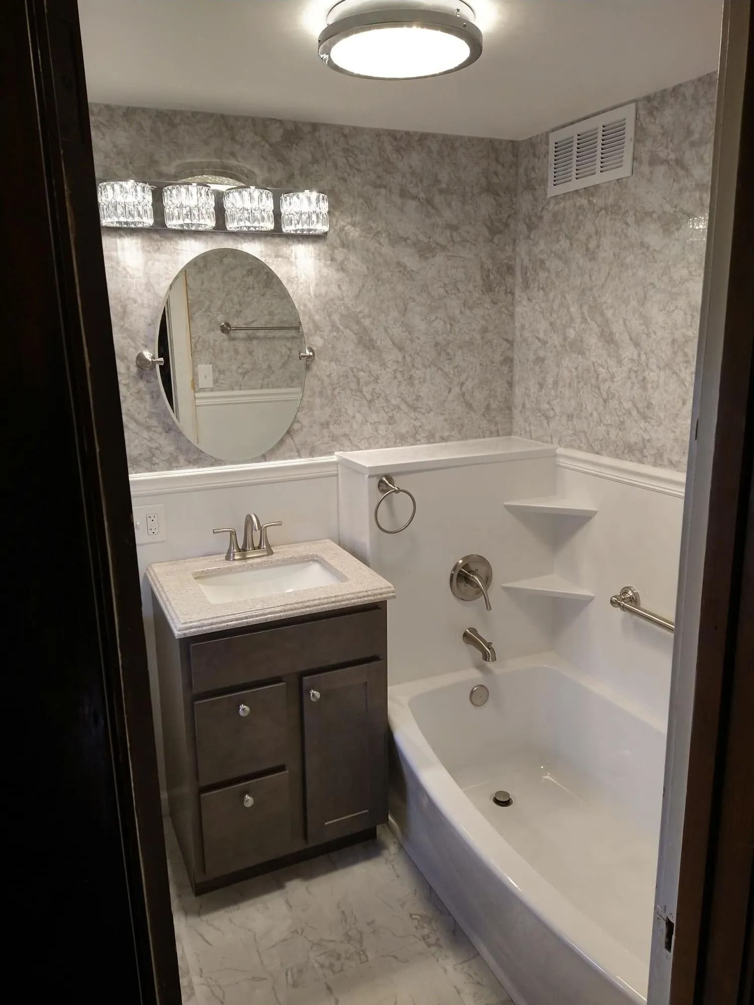A remodeled bathroom featuring a marble-patterned vanity, oval mirror, modern light fixture, and a white bathtub.