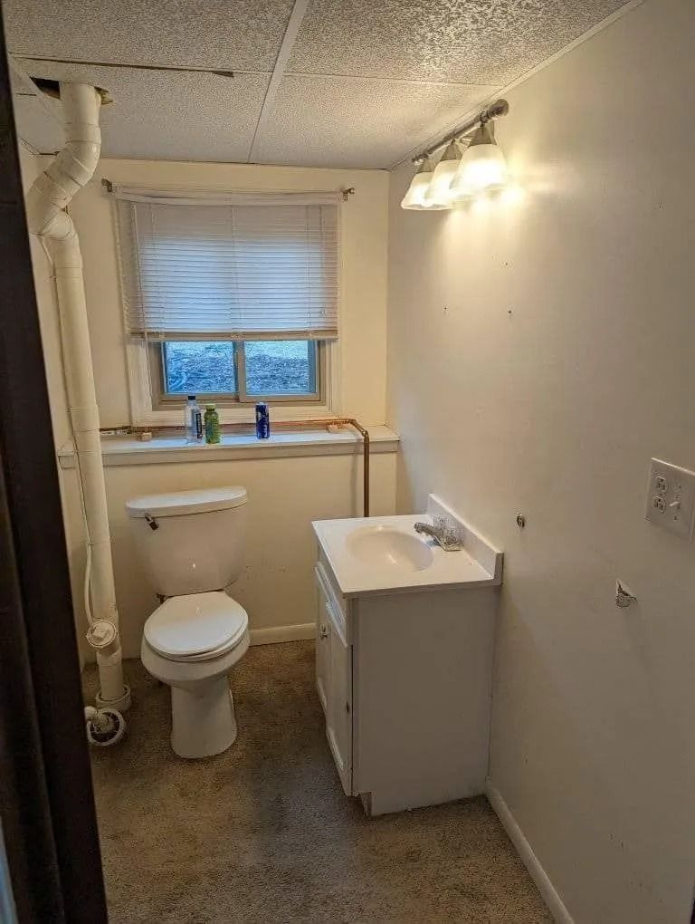 A small, windowed bathroom with a white toilet, a vanity sink, and a vertical white pipe against a cream-colored wall.