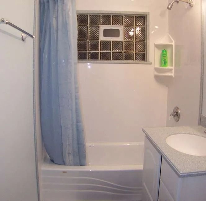 A bathroom featuring a white bathtub with a light blue shower curtain, a glass block window, and a white vanity.