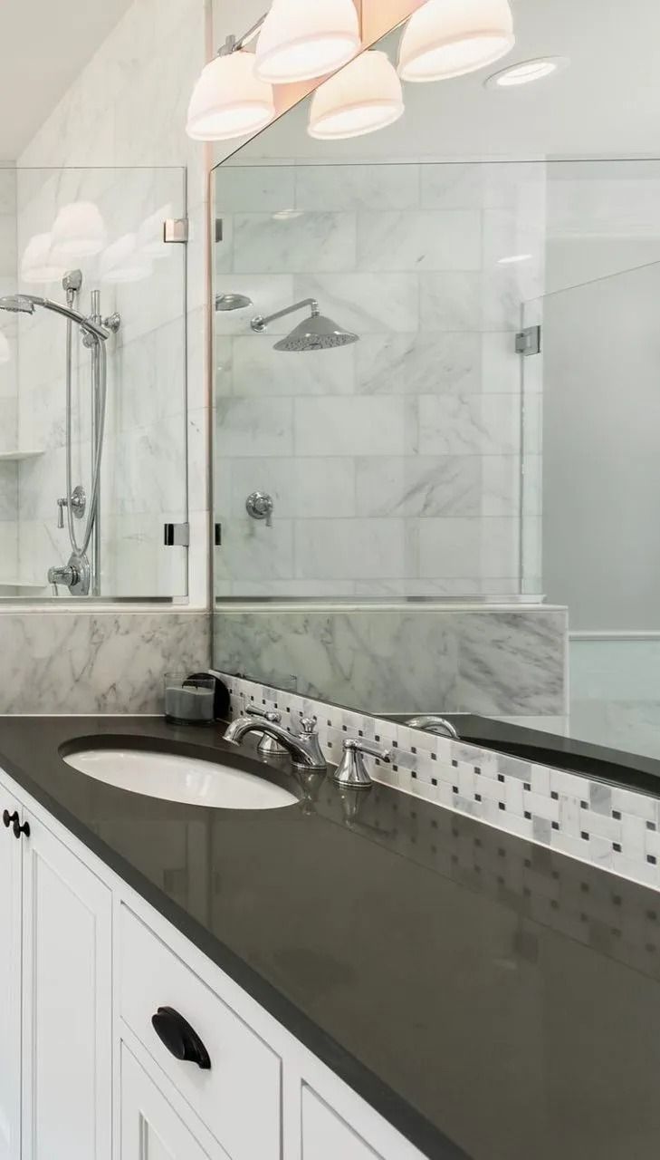 Bathroom vanity with a dark gray countertop, white cabinets, and a mosaic backsplash reflecting in a large wall mirror.