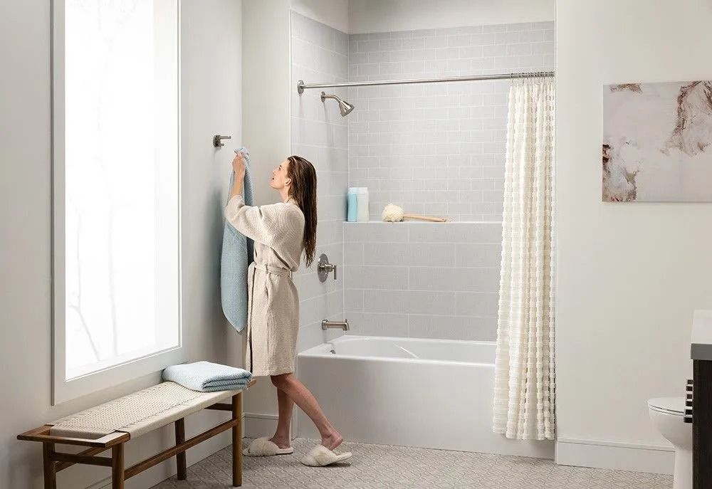 A person in a bathrobe hangs a light blue towel on a wall hook in a light-colored, modern bathroom with a bathtub.