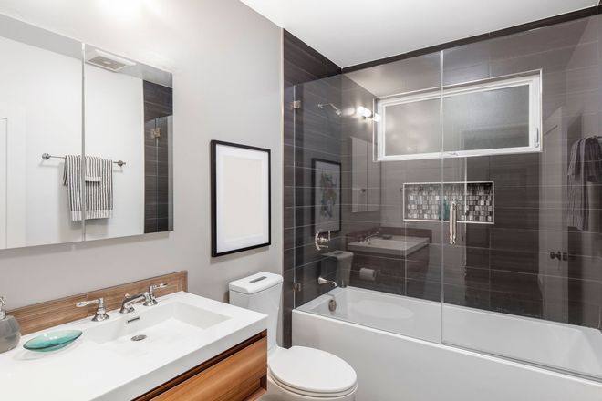 A modern bathroom featuring a white vanity with a glass sink, a toilet, and a glass-enclosed bathtub with dark tile walls.