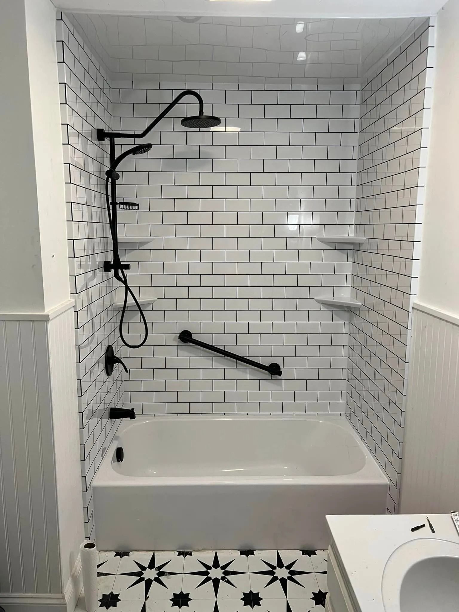 A white bathtub and shower surround with a black, geometric tile pattern, black fixtures, and a black grab bar.