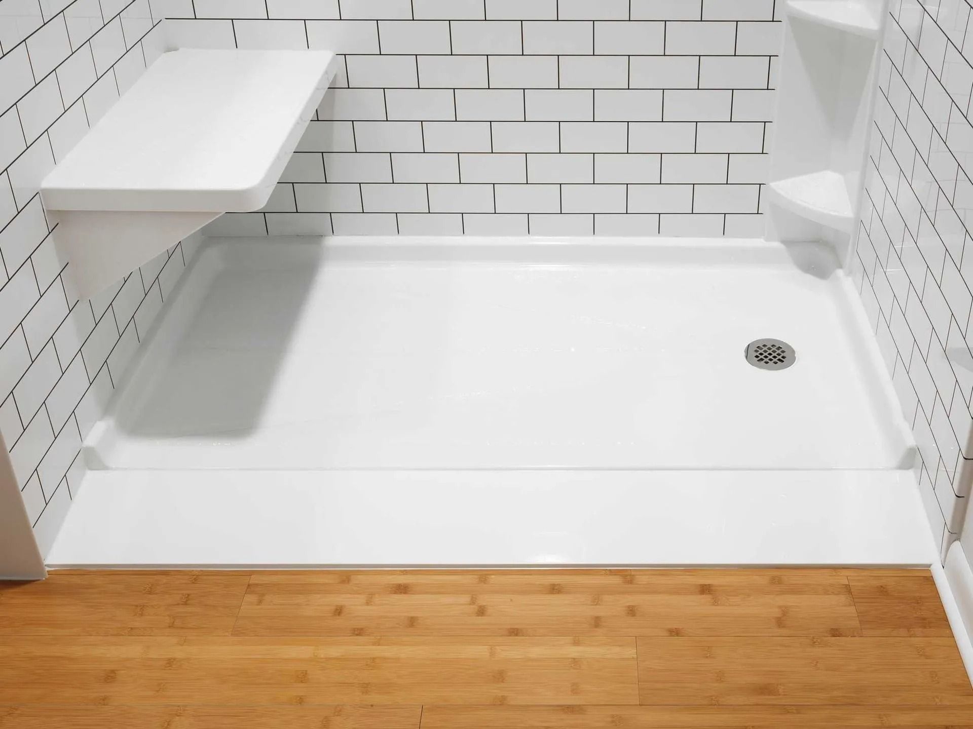 A white walk-in shower base with a built-in corner seat and shelf, set against white tiled walls and wooden flooring.