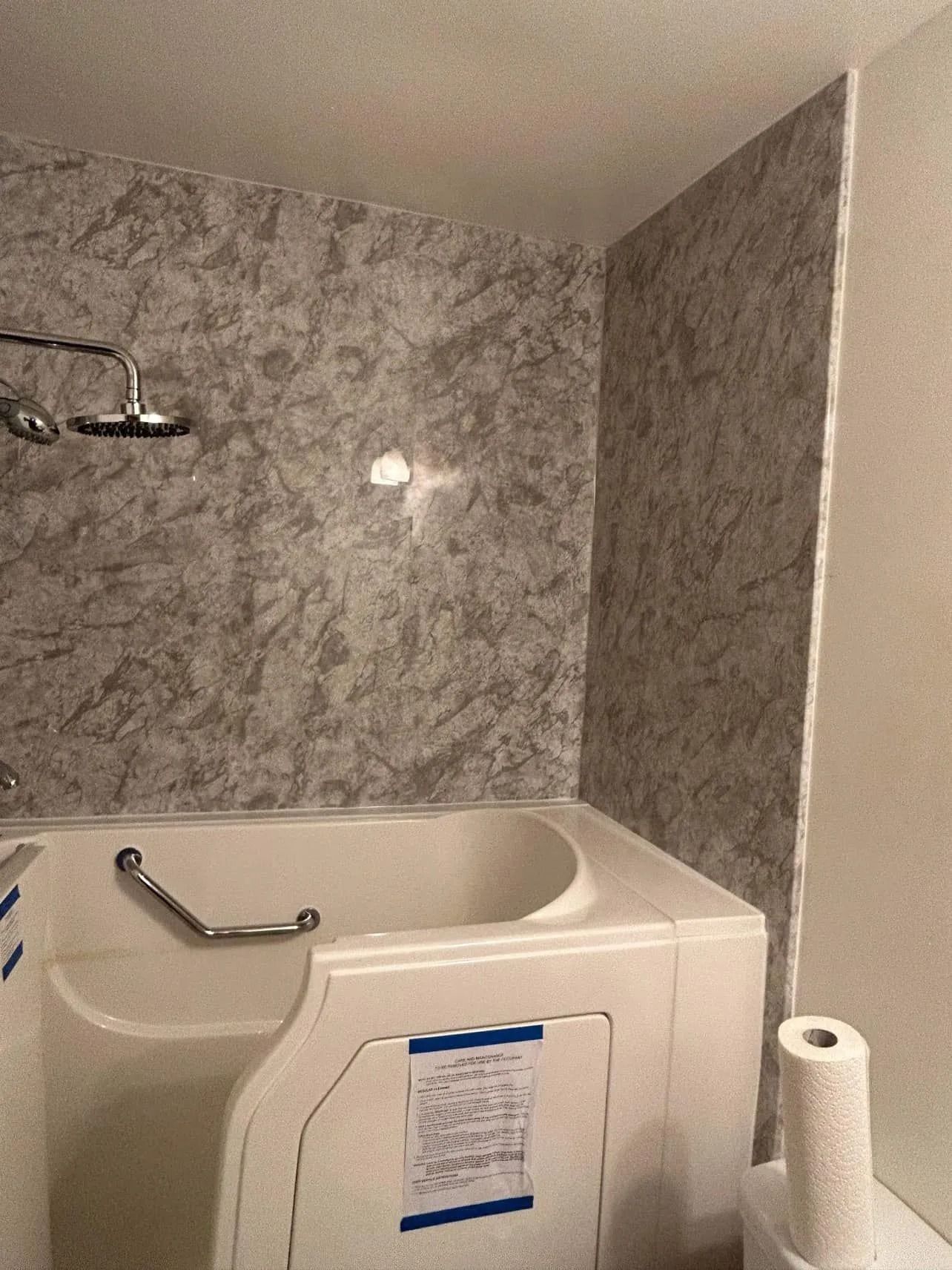Walk-in bathtub with a silver grab bar, showerhead, and gray marble-patterned wall surround in a bathroom.