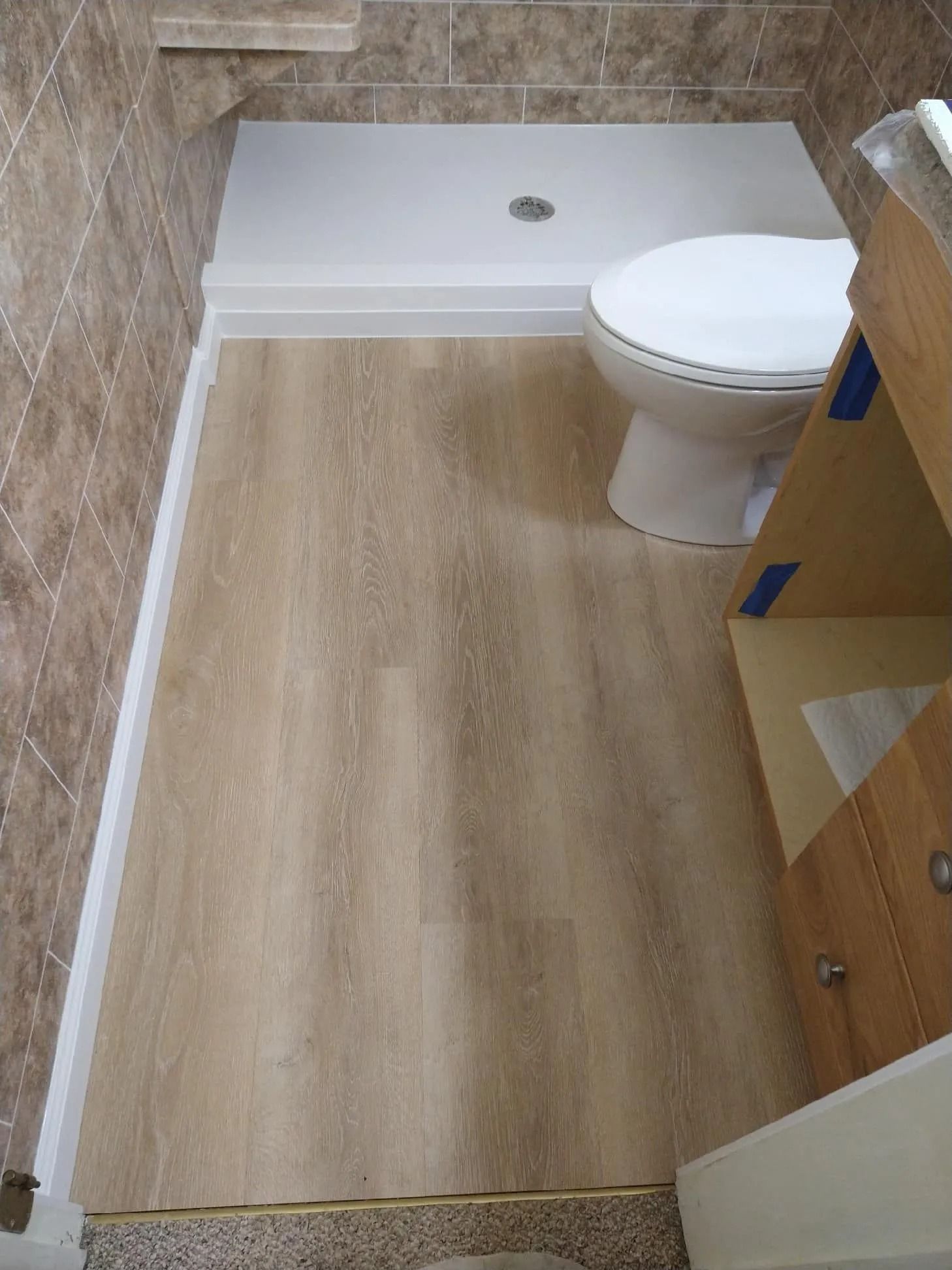 A small bathroom floor with light wood-look planks, a white shower base, and a toilet next to an unfinished vanity cabinet.