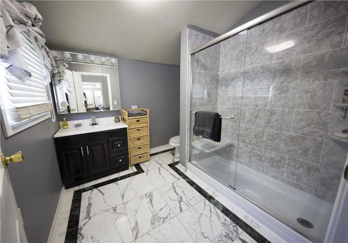 A modern bathroom with a dark vanity, white marble-style floor, and a large walk-in glass shower with stone-patterned tiles.