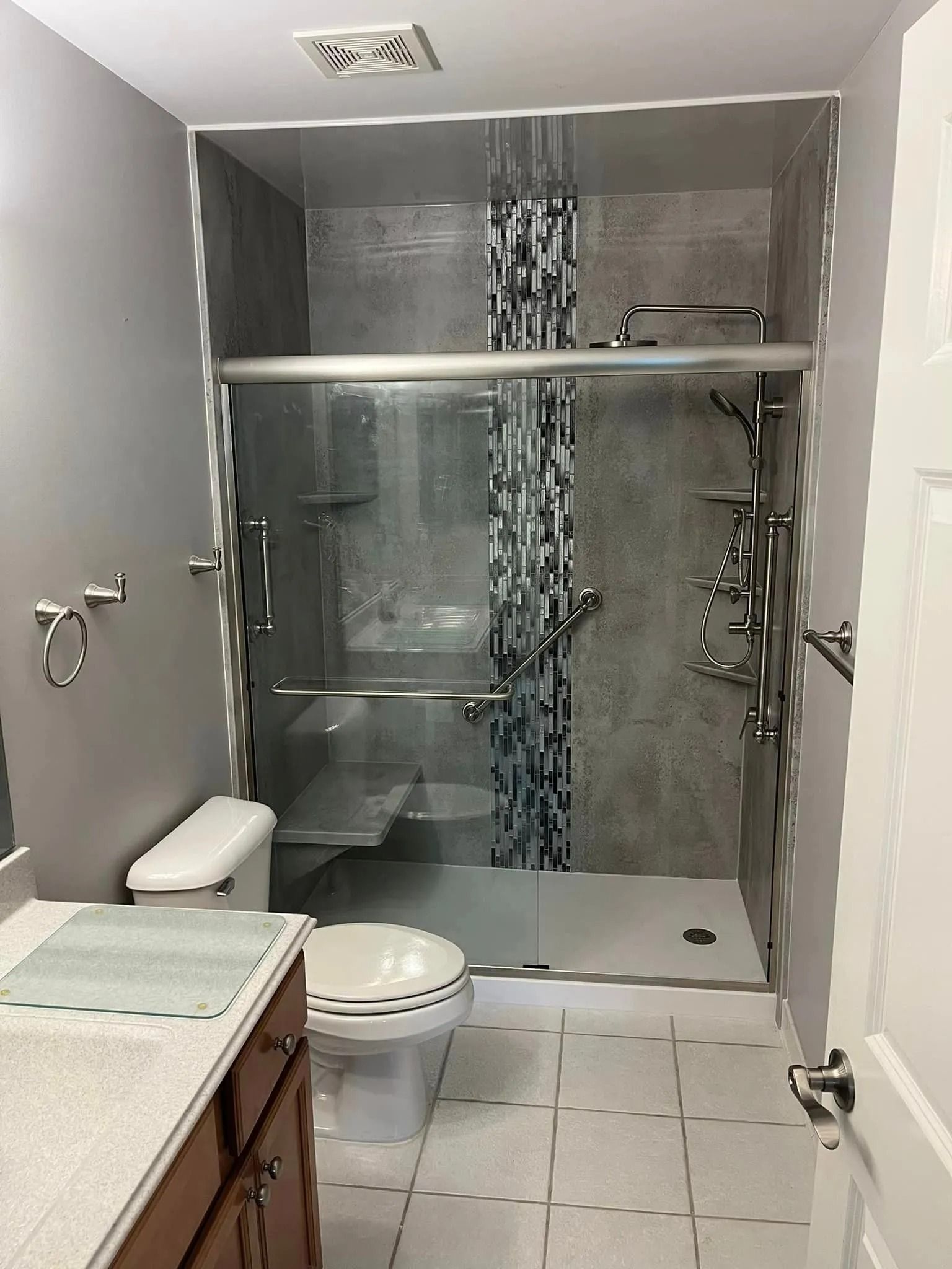 A bathroom featuring a walk-in glass shower with gray tile, a vertical mosaic accent, and a white toilet next to a vanity.