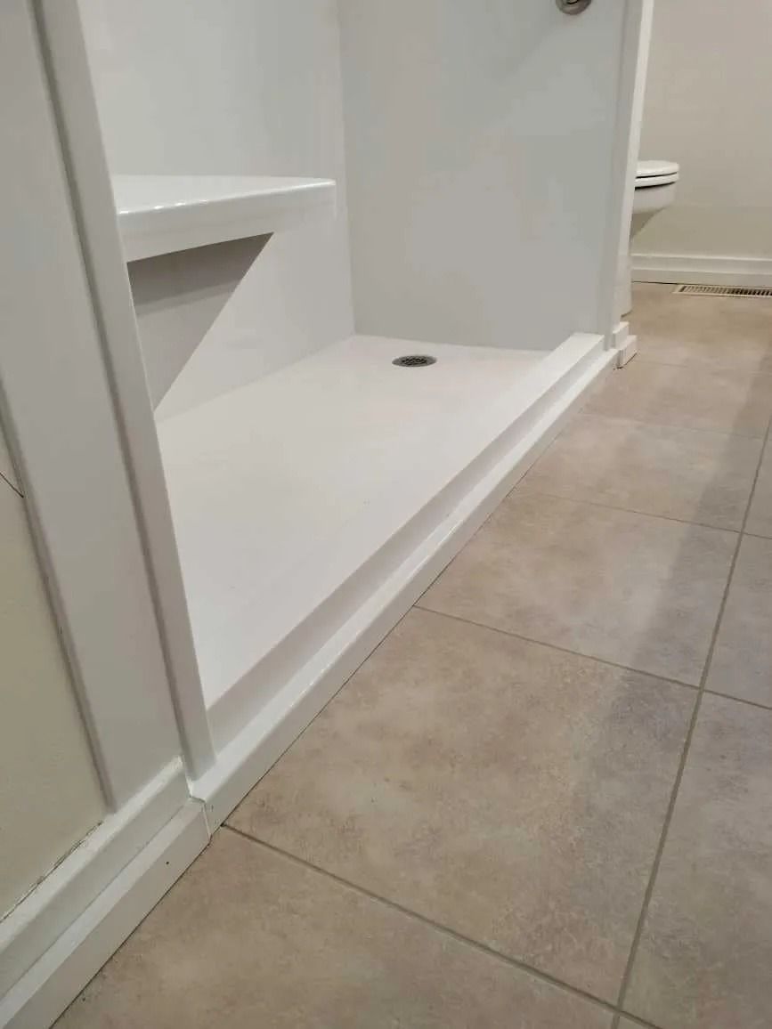 A white shower stall with a built-in seat and a low threshold, situated next to tiled bathroom flooring.